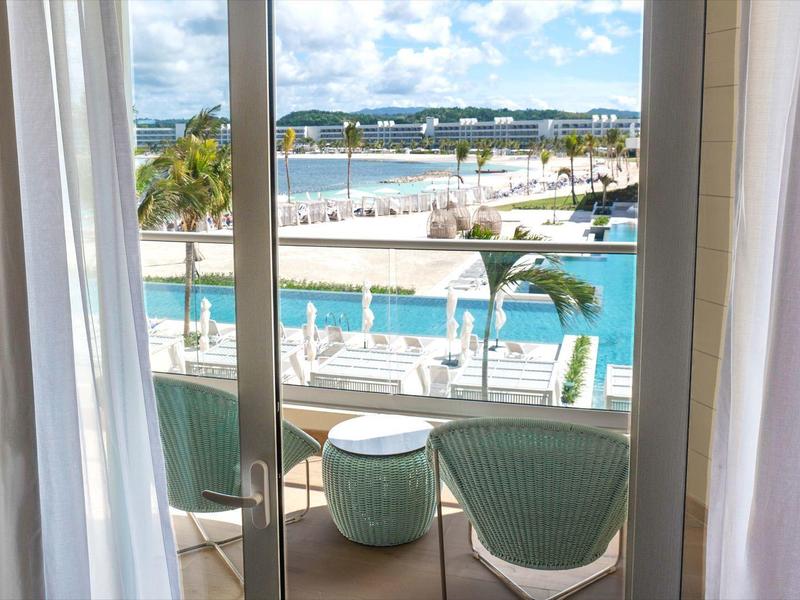 Blick durch Schiebetür auf Balkon mit grünen Sesseln und Palmen am blauen Wasser.