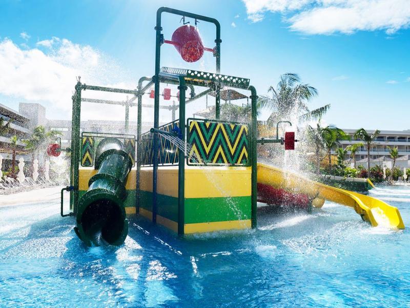 Wasserspielplatz mit Rutsche, Tunnel und bunten Elementen in einem flachen Pool bei Sonnenschein.