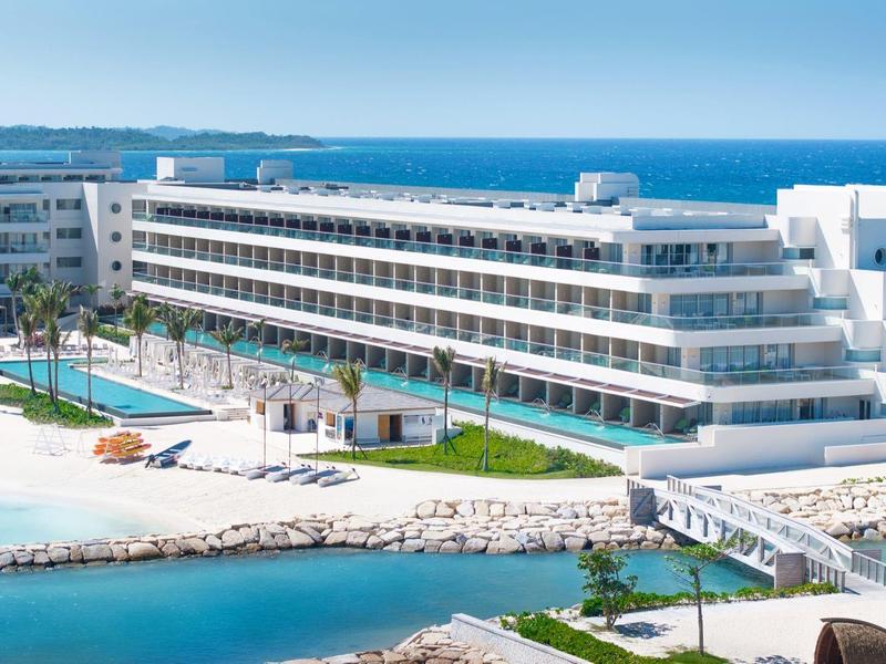 Modernes Hotelgebäude mit weißer Fassade am Wasser, Swimmingpools und Liegestühlen unter blauem Himmel.