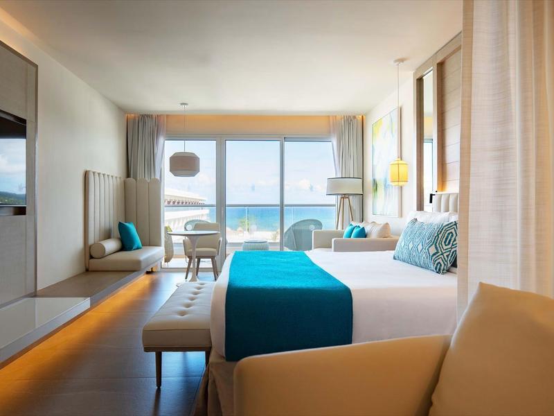 Modernes Hotelzimmer mit großem Bett, türkisfarbener Bettdecke, Meerblick und hellem Interieur.