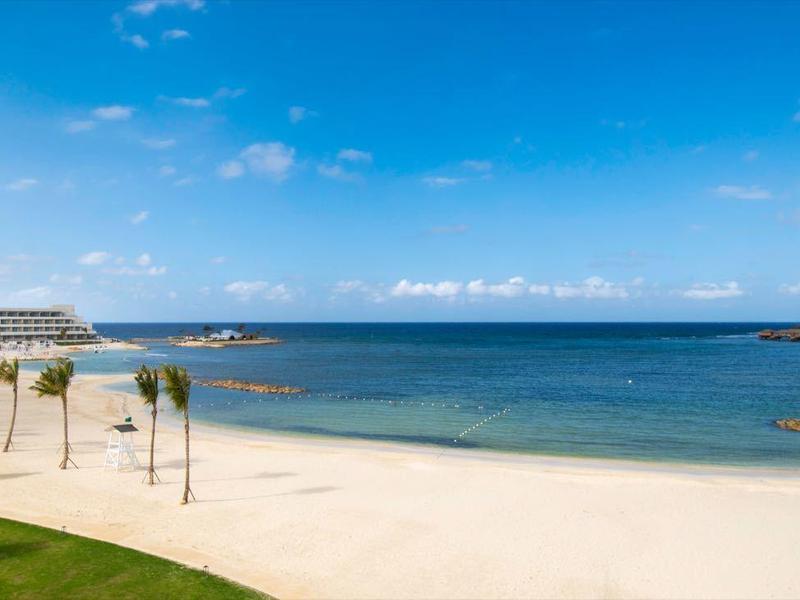 Breiter Sandstrand mit Palmen, blauem Meer und klarem Himmel, links weiße Gebäude und Grünflächen.