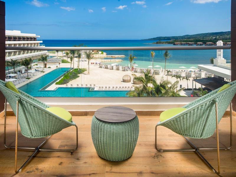 Zwei grüne Stühle und ein kleiner Tisch auf Balkon mit Blick auf Pool, Strand und blaues Meer.