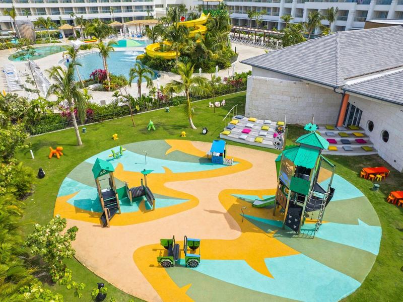 Bunter Spielplatz mit Rutsche, Schaukeln und Kletterturm umgeben von Gras und Gebäuden.