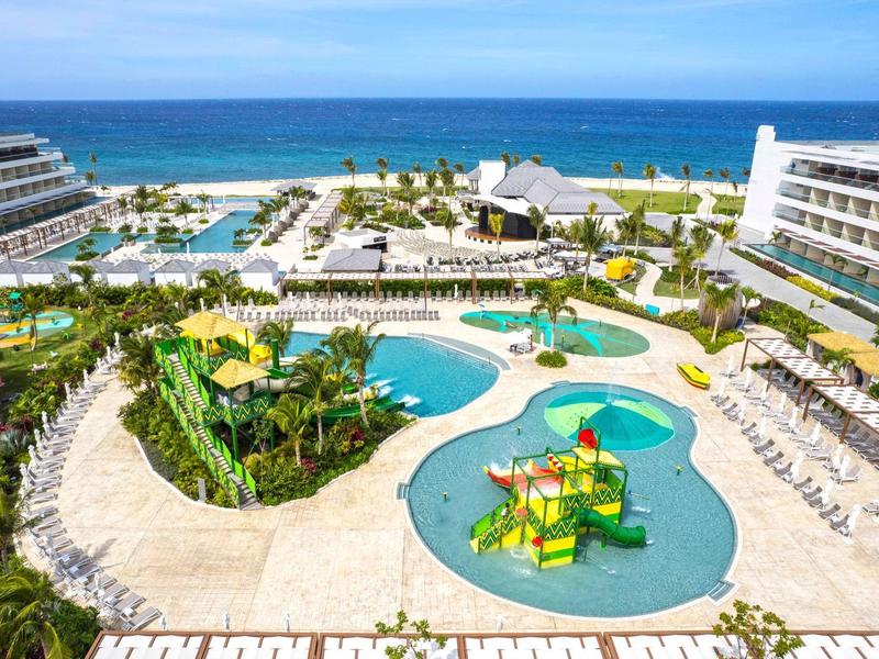 Große Resortanlage mit Pools, Wasserspielplatz, Palmen und Blick auf das blaue Meer im Hintergrund.