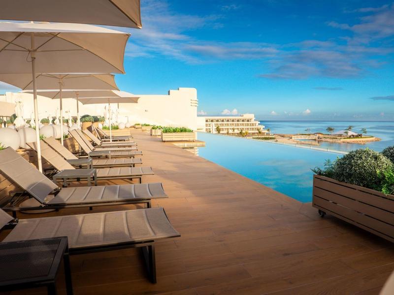 Lounge-Sessel am Pool mit Holzterrasse, Sonnenschirmen und blauem Himmel im Hintergrund.