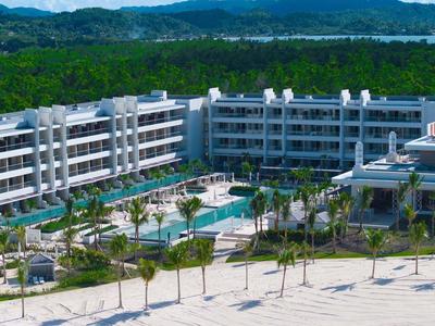 Large modern beach resort with pool and palm trees in front of wooded hills.