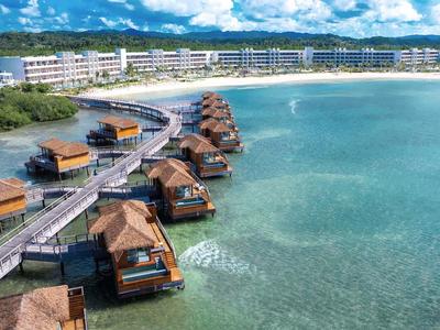 Overwater bungalows on clear turquoise water beach with a hotel in the background.