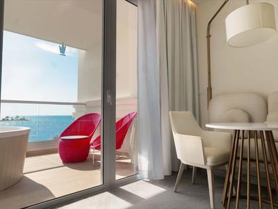 Bright living area overlooking balcony with bathtub and red seating by the sea.