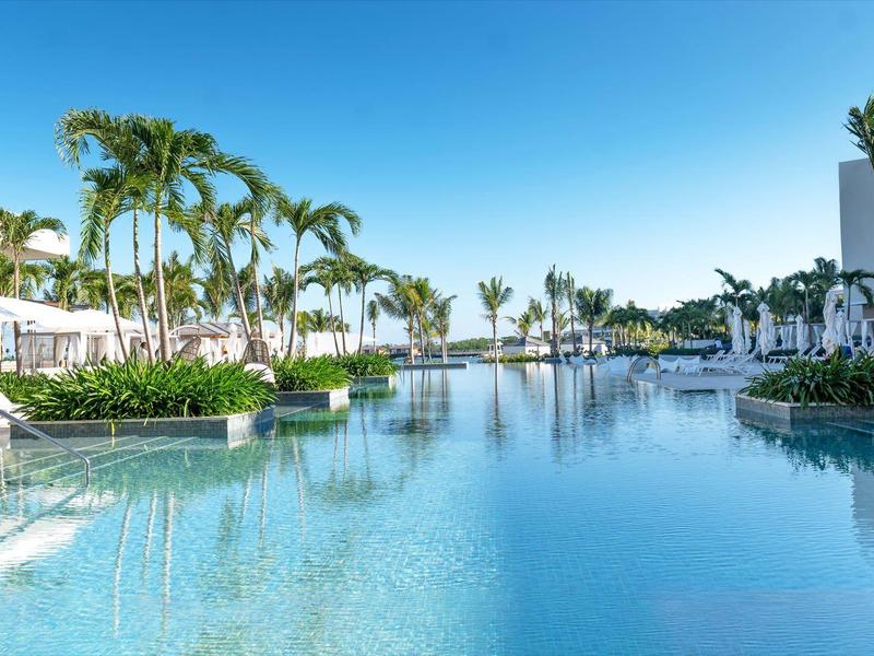 Large pool with palm trees and umbrellas at a modern hotel resort under clear sky.