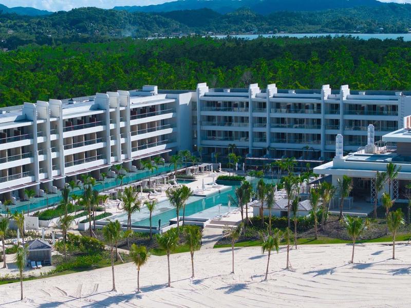 Large modern beach resort with pool and palm trees in front of wooded hills.