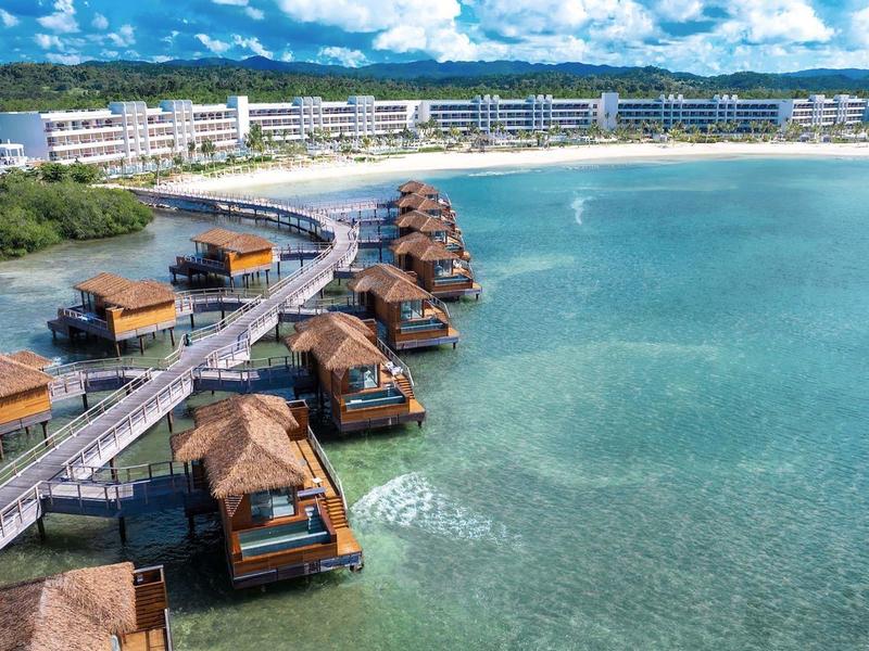 Overwater bungalows on clear turquoise water beach with a hotel in the background.
