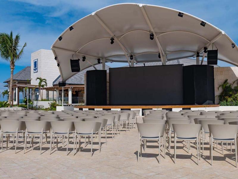 Large stage with screen and rows of chairs on the beach under a blue sky.