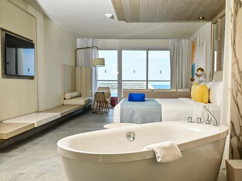 Modern hotel room with freestanding bathtub and sea view through large windows.