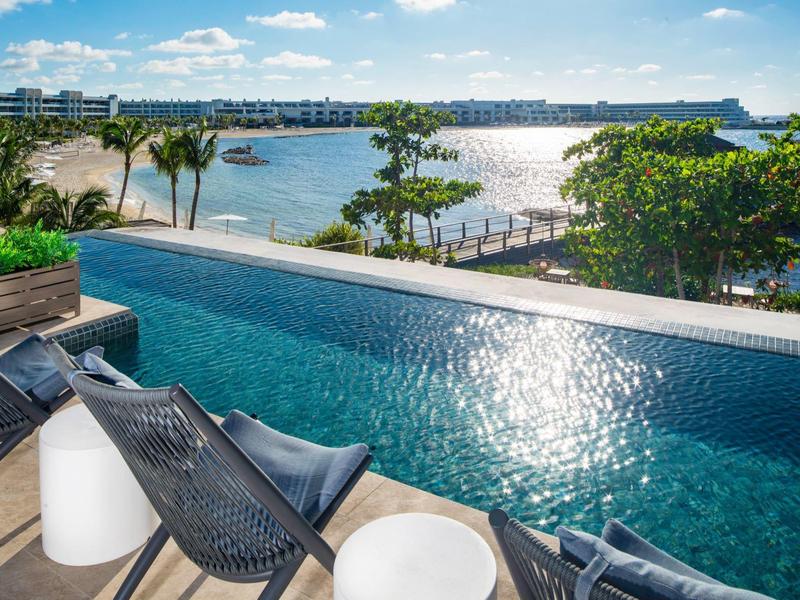 View of a pool with chairs by the water, palm trees, and cityscape in the background in sunlight.