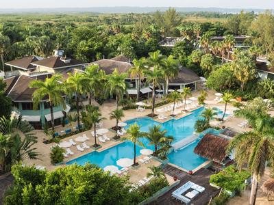 Hotel mit mehreren Pools umgeben von Palmen und grüner Landschaft unter wolkenlosem Himmel.