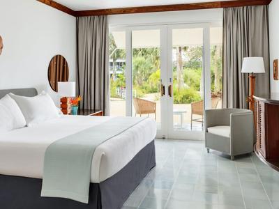Helles Hotelzimmer mit Doppelbett, Sesseln und Balkon mit Blick auf Garten.