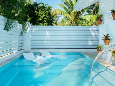 Kleiner Pool mit klar blauem Wasser, weißen Liegen und Pflanzen an einer weißen Holzwand.