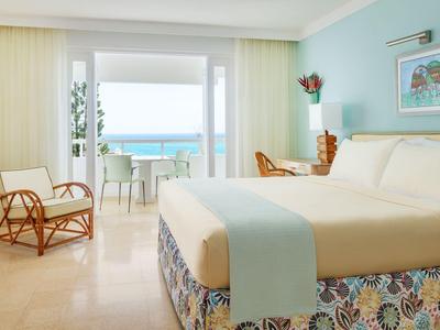 Helles Hotelzimmer mit Doppelbett, Rattanstuhl und Balkon mit Meerblick.