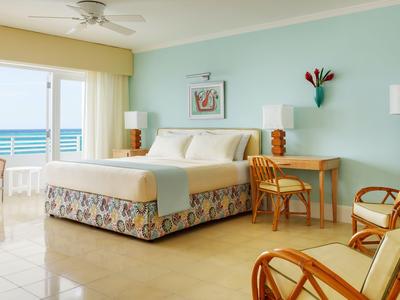 Habitación de hotel luminosa con cama grande, muebles de ratán y vista al mar por grandes ventanas.
