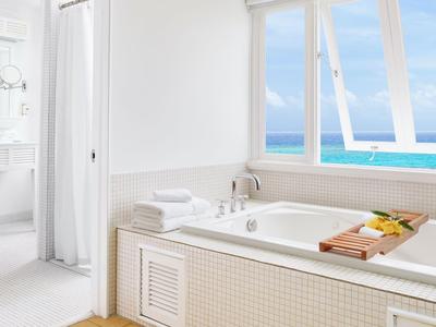 Baño luminoso con bañera, decoración blanca y vista al mar a través de ventanas abiertas.