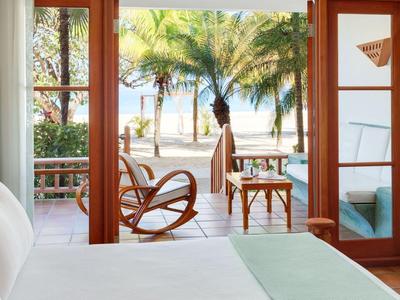 Blick von einem Hotelzimmer auf eine Terrasse mit Stühlen und Palmen zum Strand hin.