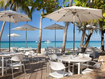 Terrasse mit weißen Tischen und Stühlen unter Sonnenschirmen am Strand mit Palmen.