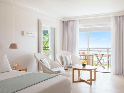 Helles Hotelzimmer mit Bett, Sesseln und Balkon mit Blick auf das Meer.