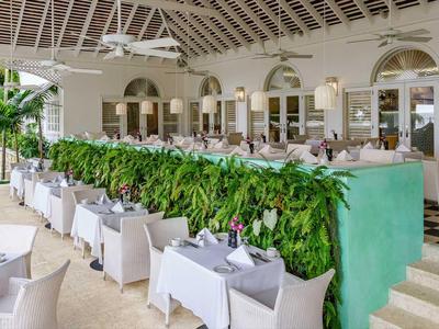 Großer Hotelrestaurantbereich mit weißen Tischen und Stühlen, Grünpflanzen als Raumteiler und offenem Dach.