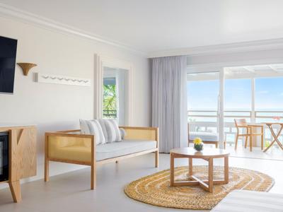Helles Hotelzimmer mit Holzmöbeln, Blick auf Balkon und Meer mit blauem Himmel.
