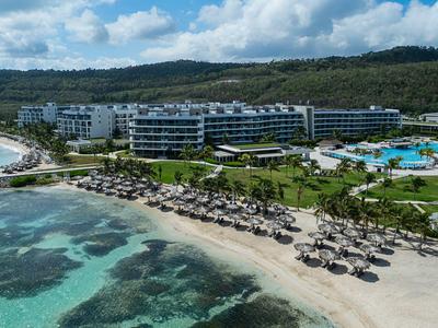 Luxushotel mit Pools am Strand und klarem Meerwasser, umgeben von grünen Hügeln.