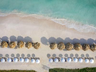 Vogelperspektive auf einen Sandstrand mit Reihen von besonnten Sonnenschirmen und türkisfarbenem Wasser.