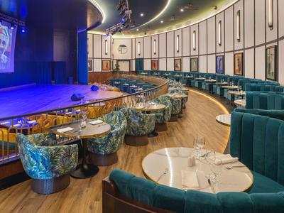 Eleganter Veranstaltungsraum mit Bühne, runden Tischen und grünen Sesseln in einem Hotel.