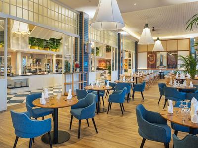 Helles Restaurant mit blau gepolsterten Stühlen, runden Holztischen und modernen Pendelleuchten.