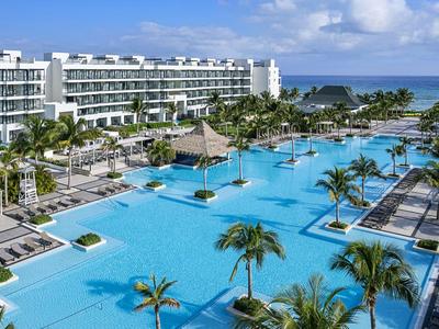 Großer Hotelpool mit Palmen, Liegestühlen und Blick aufs Meer an einem sonnigen Tag.