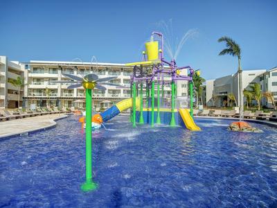 Großer Außenpool mit Wasserrutsche und Spielgeräten vor einem modernen Hotelgebäude.