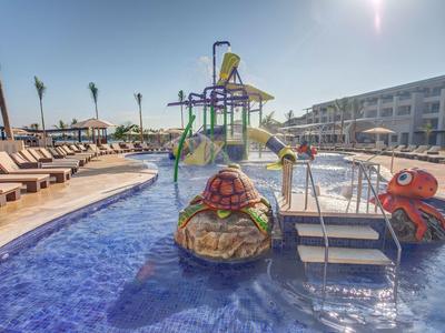 Hotelpool mit Wasserspielplatz und Liegestühlen unter klarem Himmel.