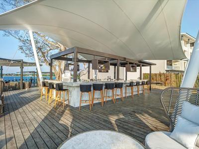 Moderner Strandbarbereich mit weißem Segel über Holzterrasse und Bar mit Hockern.