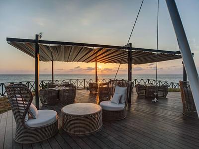 Gemütlicher Außenbereich mit Rundbänken und Tischen auf Holzdeck mit Blick auf Sonnenuntergang am Meer.