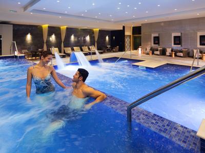 Zwei Menschen entspannen im beleuchteten Innenpool eines Hotels mit Liegestühlen im Hintergrund.