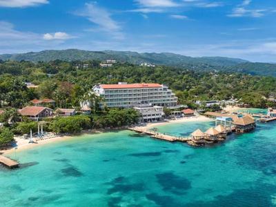 Luchtfoto van een hotelresort aan de turquoise zee met groen kustlandschap en bootsteigers.