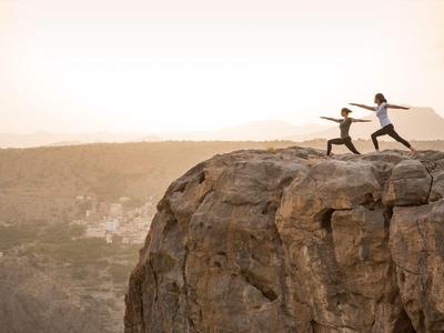 Zwei Personen machen Yoga-Übungen auf einem Felsen mit Aussicht auf ein Tal bei Sonnenaufgang.