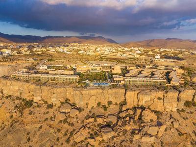 Desert landscape with rocky cliffs, scattered buildings, and distant mountains under a cloudy sky.