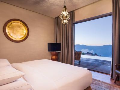 Cozy hotel room with bed, warm lighting, and balcony overlooking mountains and clear sky.