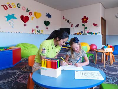 Child and teacher drawing together at a blue table in a colorful playroom.