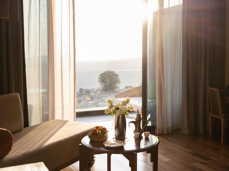 Sunlit hotel room with a chaise lounge, small table, and sea view through sheer curtains.