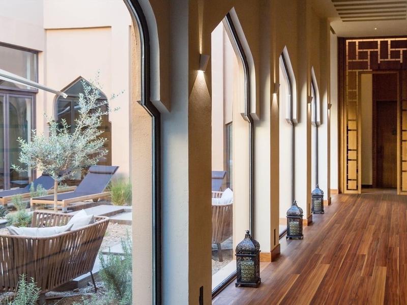 Arched corridor with wooden floor and lanterns overlooking a courtyard with wicker chairs and plants.