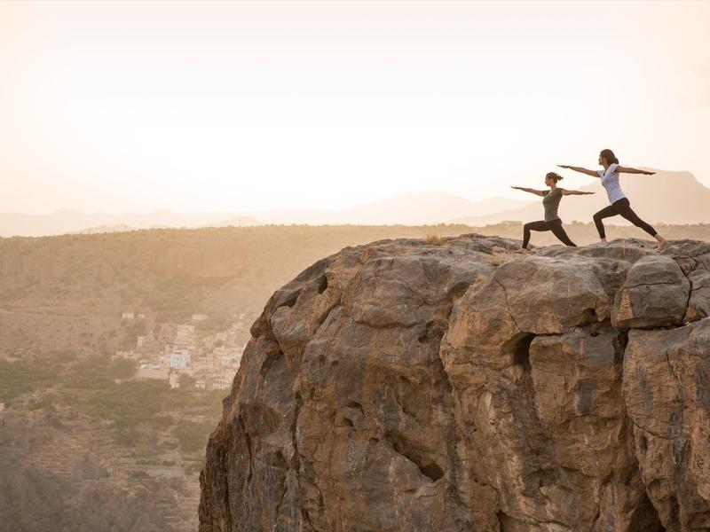 Two people practicing yoga on a rocky cliff at sunset with a scenic view below.