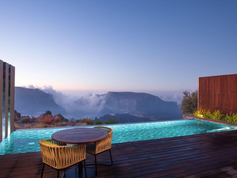 Infinity pool overlooking misty mountains at sunrise with wooden deck and table with chairs.