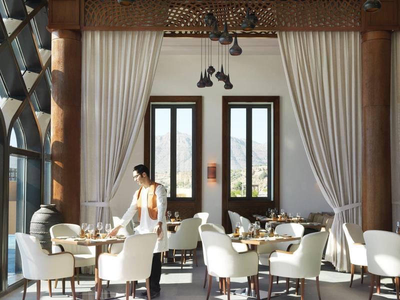 Bright hotel dining area with white chairs, wooden columns, and large windows with curtains.
