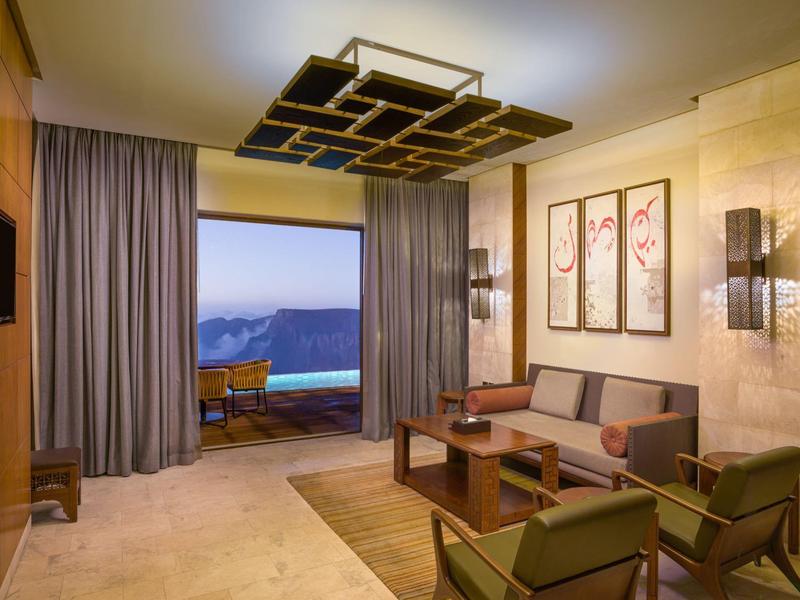 Luxurious hotel room with modern furniture and a balcony overlooking mountains at dusk.